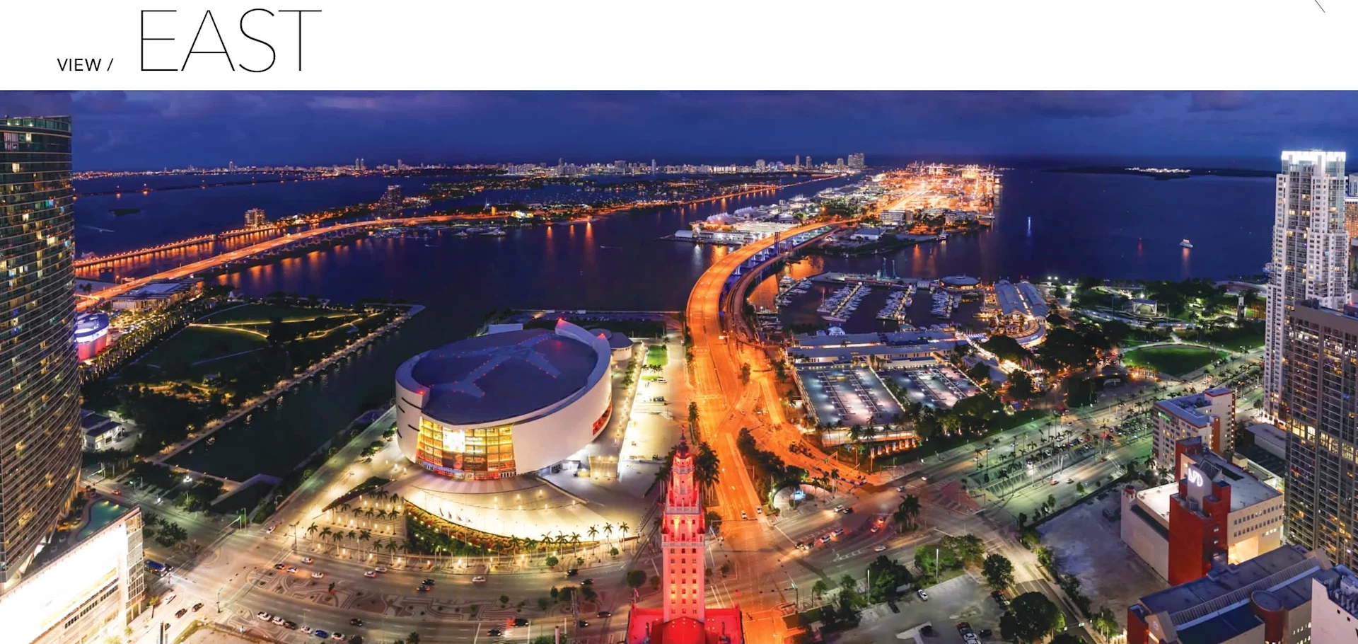 Aerial view of a vibrant cityscape at night, featuring illuminated buildings, a waterfront, and a winding road.