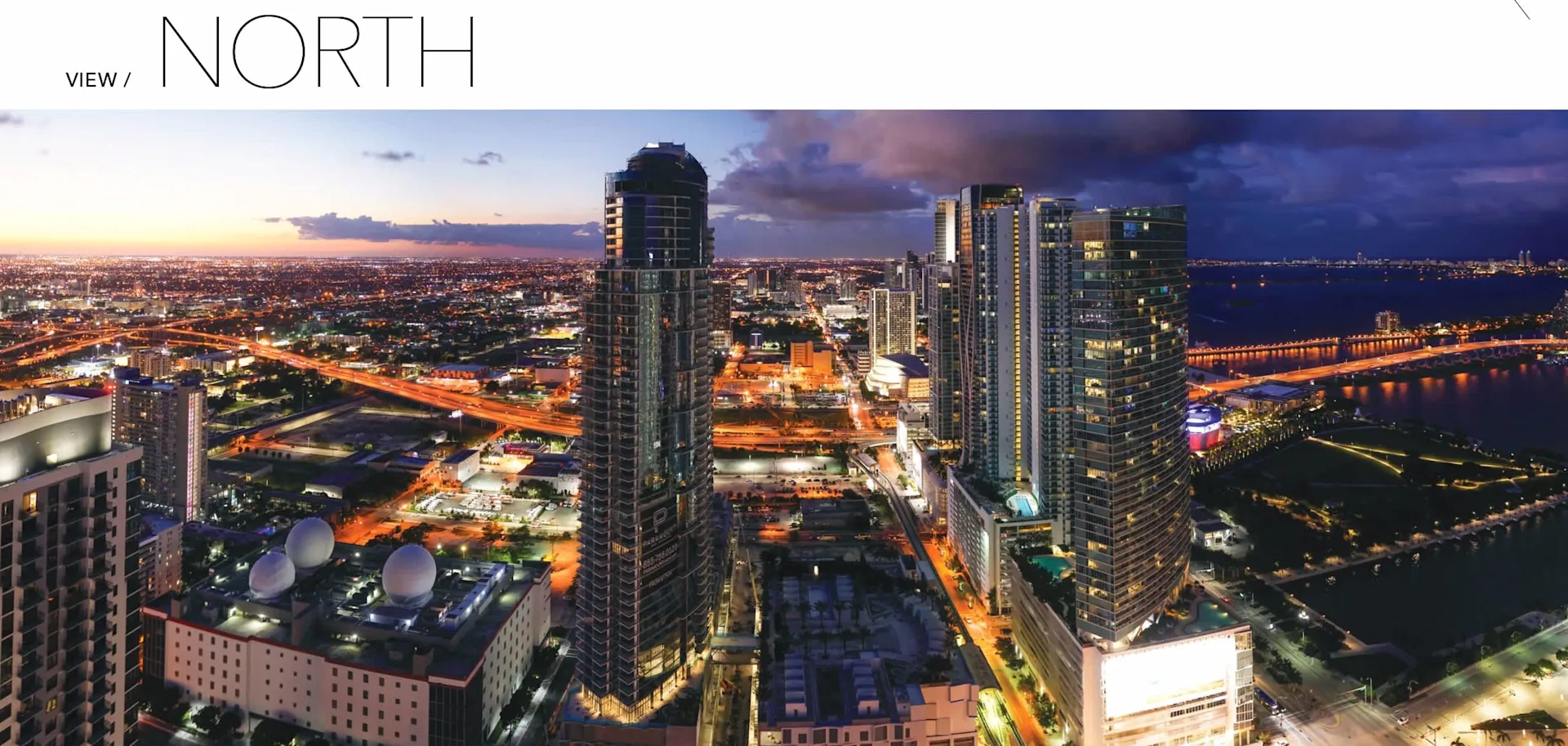 A panoramic view of a city skyline at dusk, featuring tall buildings, illuminated streets, and a river. The sky transitions from blue to orange.