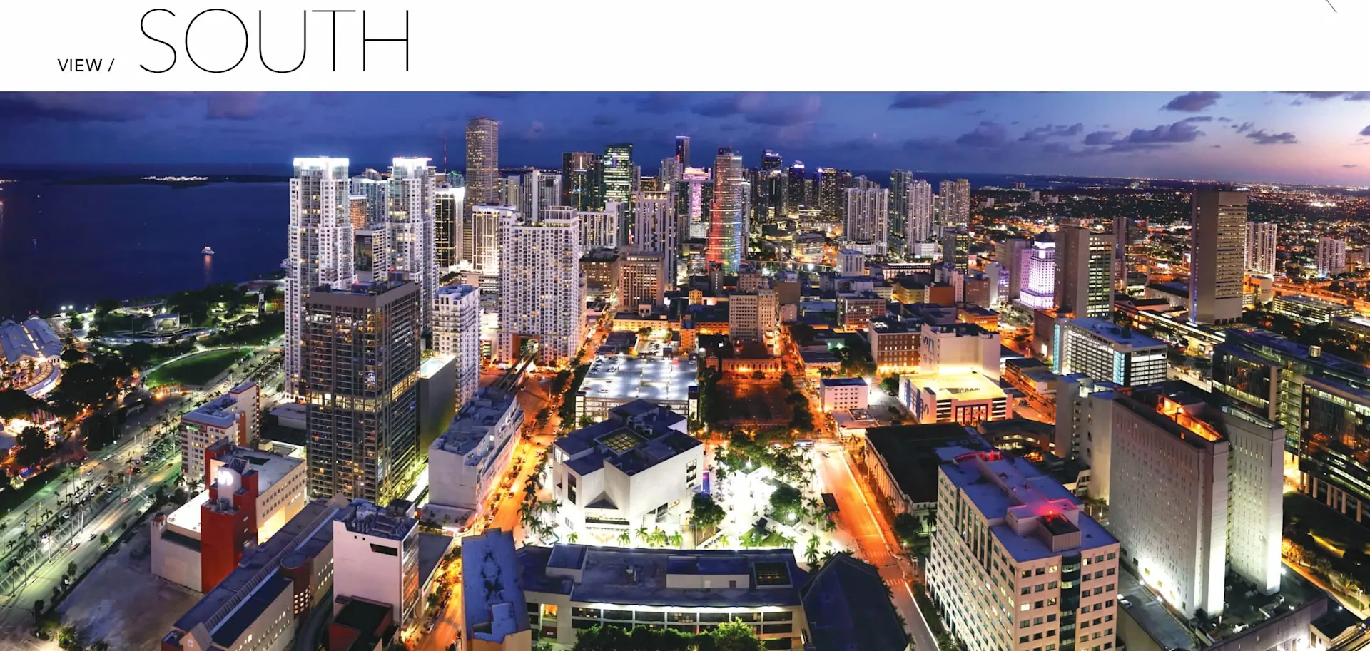 Aerial view of a vibrant city skyline at night, showcasing illuminated buildings and streets, with a body of water in the background.